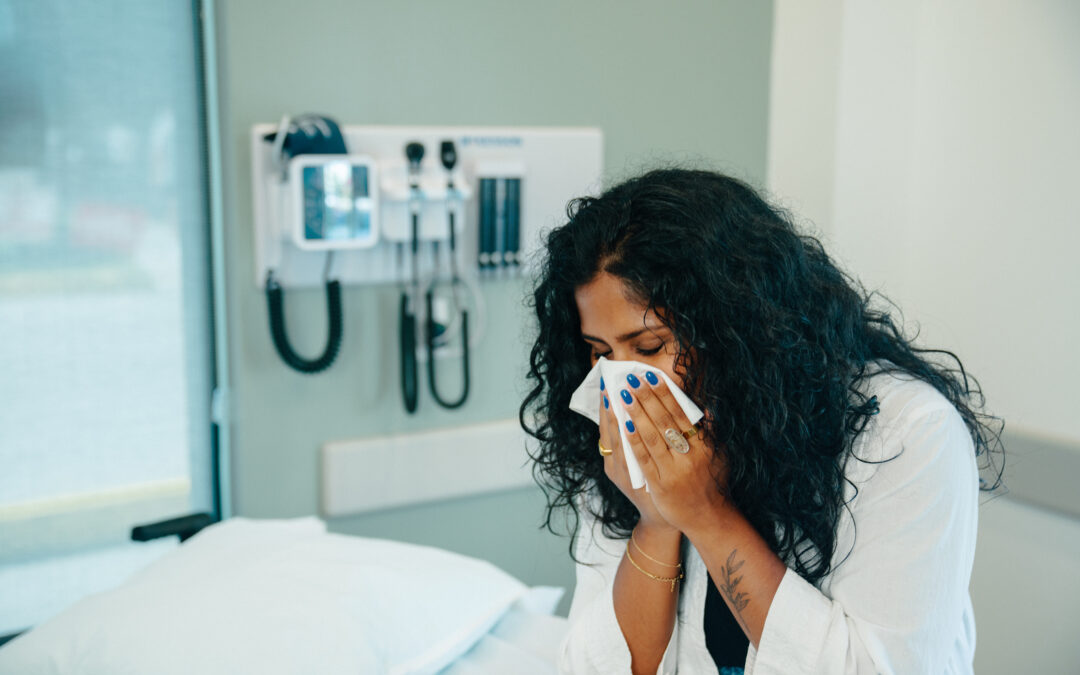 Woman with cold sitting in urgent care clinic examination room, blowing her nose or sneezing into facial tissue, with medical instruments behind her on wall.