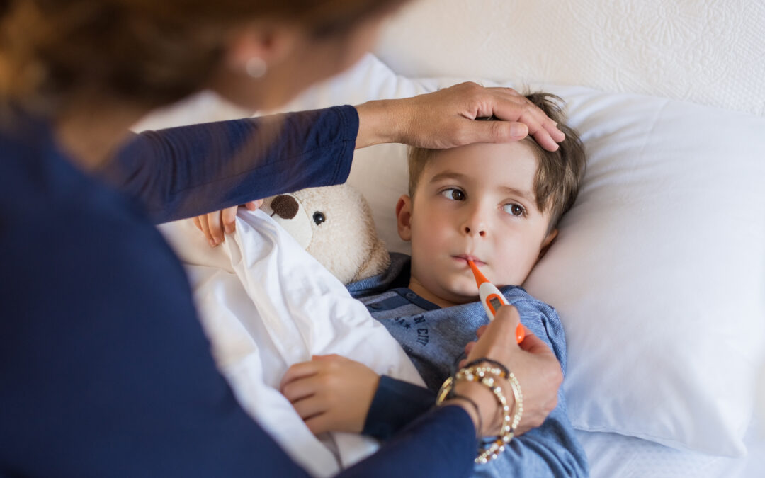 Woman checking young boy's temperature with thermometer while the boy lies in bed hugging stuffed animal. Women also checks boy’s forehead with her hand to see if he's running a fever.